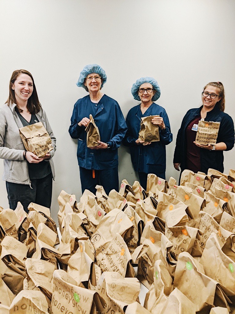 Four women standing behind a large number of brown paper bags holding up brown paper bags