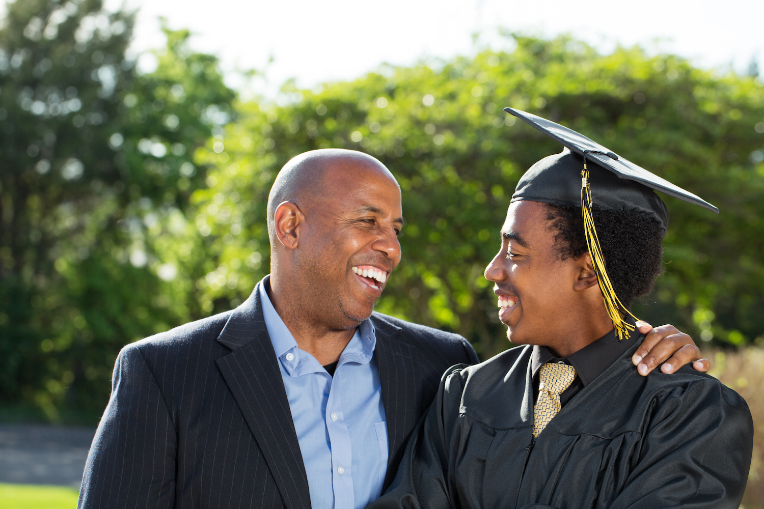 person in a blazer and button down shirt with their arm around a younger person (presumably their son) wearing a graduation cap and gown
