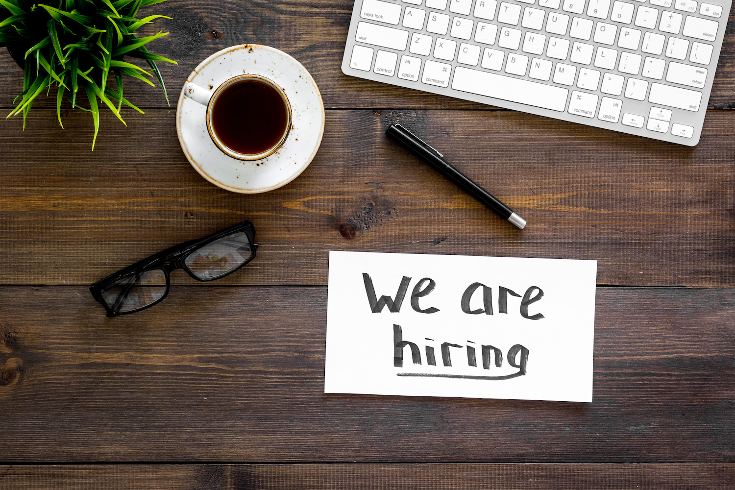 aerial view of a desk with eye galsses, a coffe,cup, a notepad that says we are hiring on it