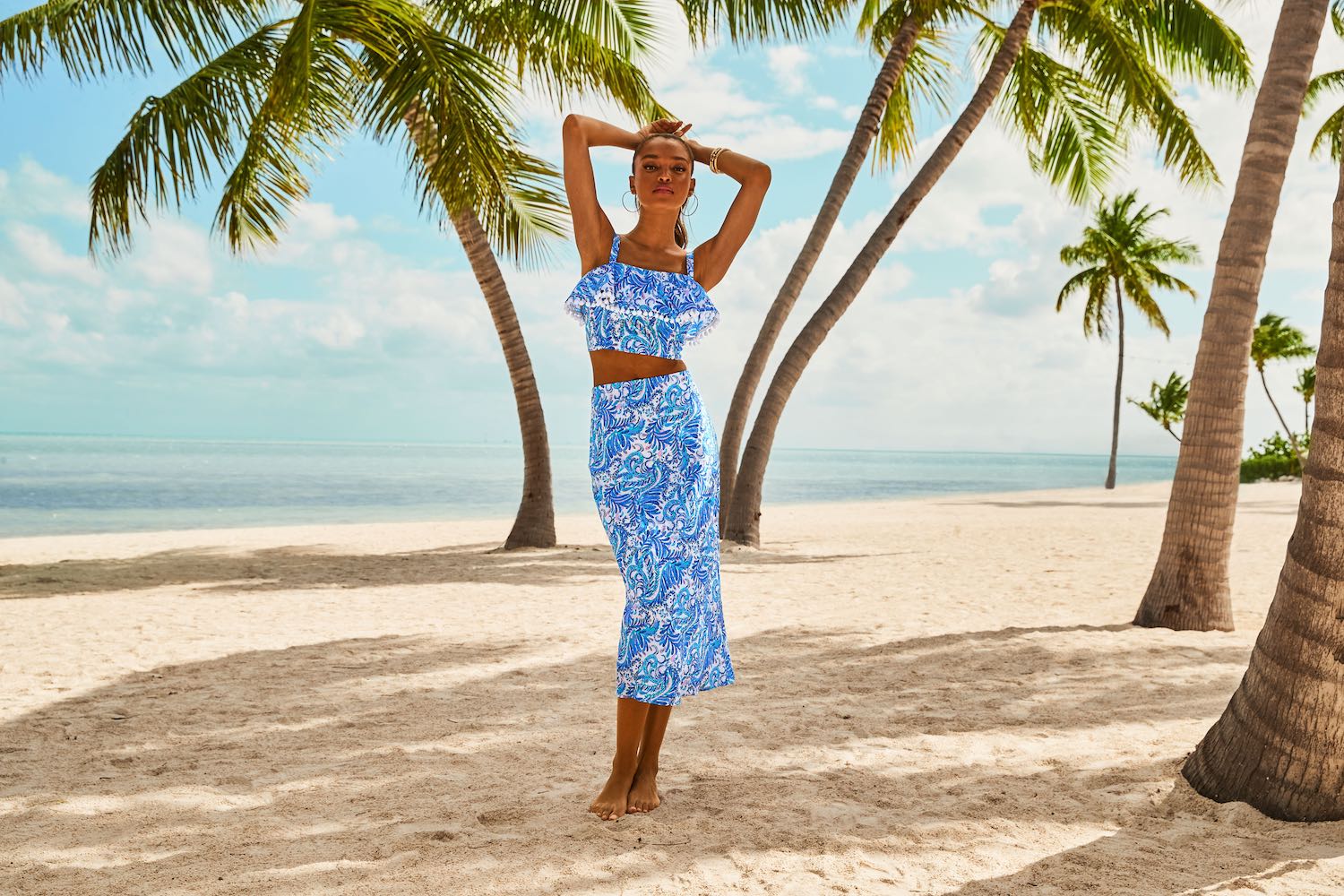 person in brightly colored crop top and long skirt standing on a beach with palm trees in the background
