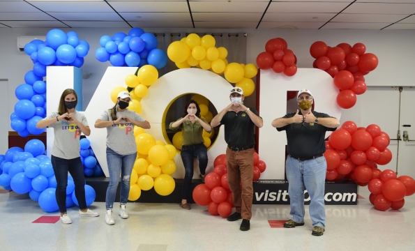 five people standing in front over oversized K-O-P letters that are covered in blue, yellow and red balloons making hearts with their hands