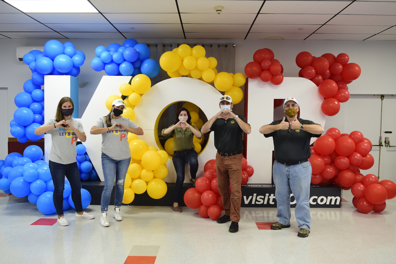 five people standing in front over oversized K-O-P letters that are covered in blue, yellow and red balloons making hearts with their hands
