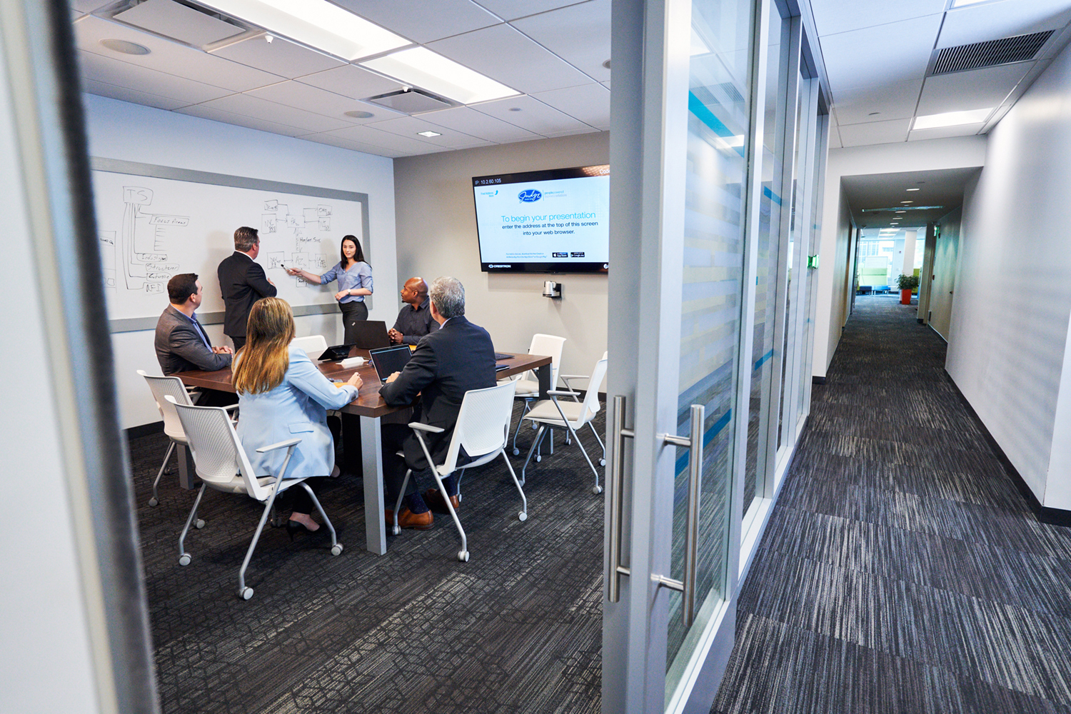 inside of a room with a glass door with people seated at a table while another person points to a white board