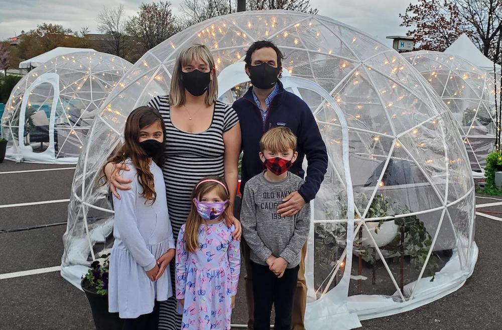 family standing outside a large clear plastic dining igloo wearing masks