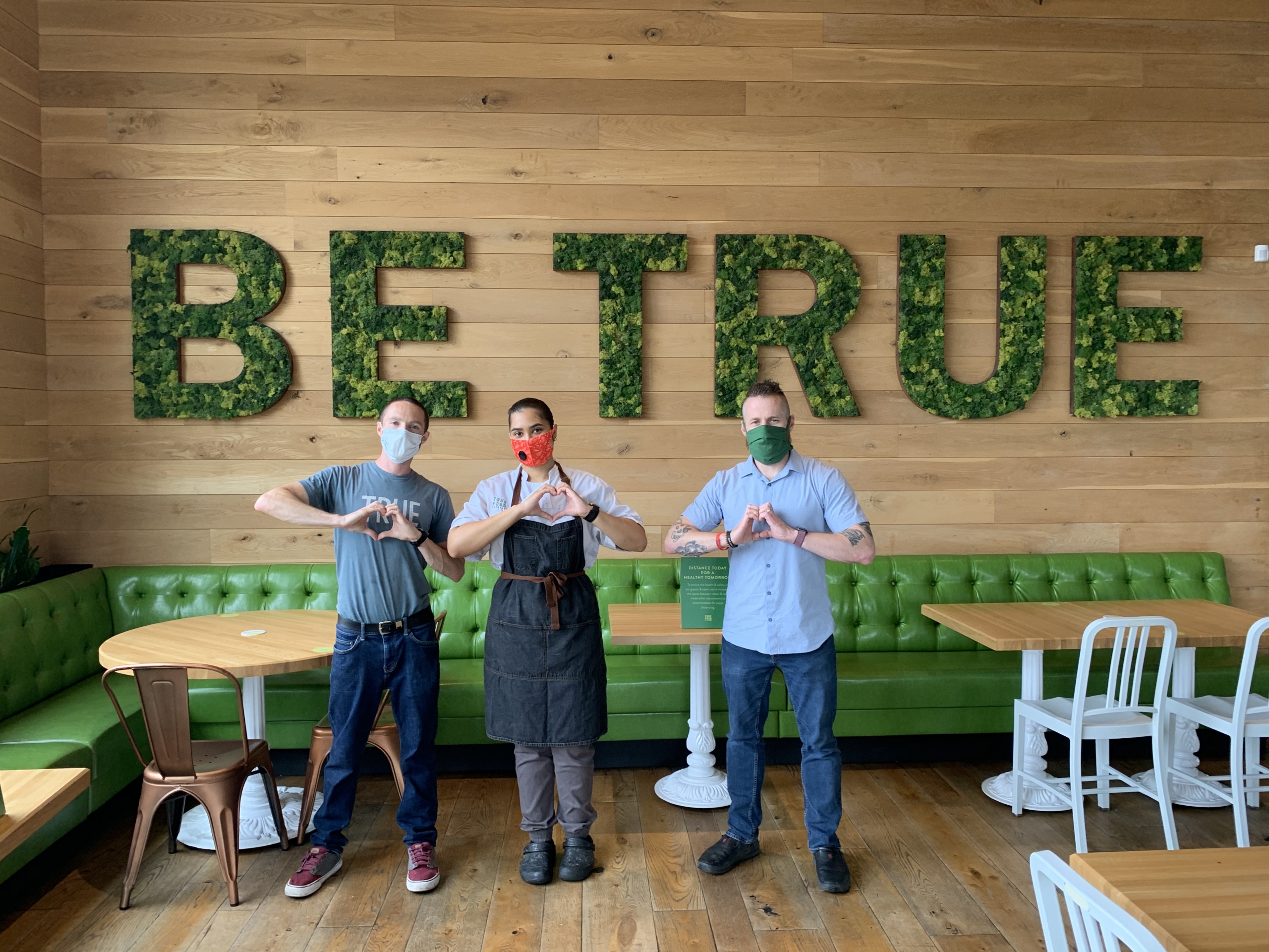 three people in front of a large wall that says "BE TRUE" on it, making heart symbols with their hands