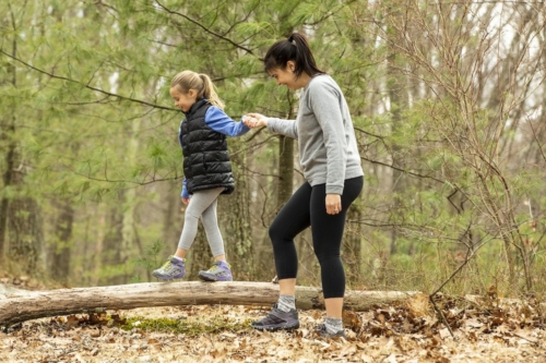 person outdoors in the woods holding a childs hand as they walk across a log