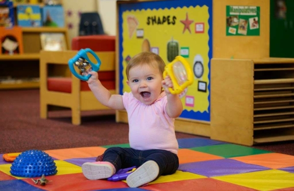 baby sitting on colorflul floor mat, holding up toys and smiling