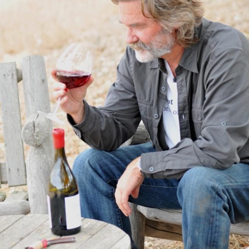 Kurt Russel sitting on a chair outdoors with a glass of red wine