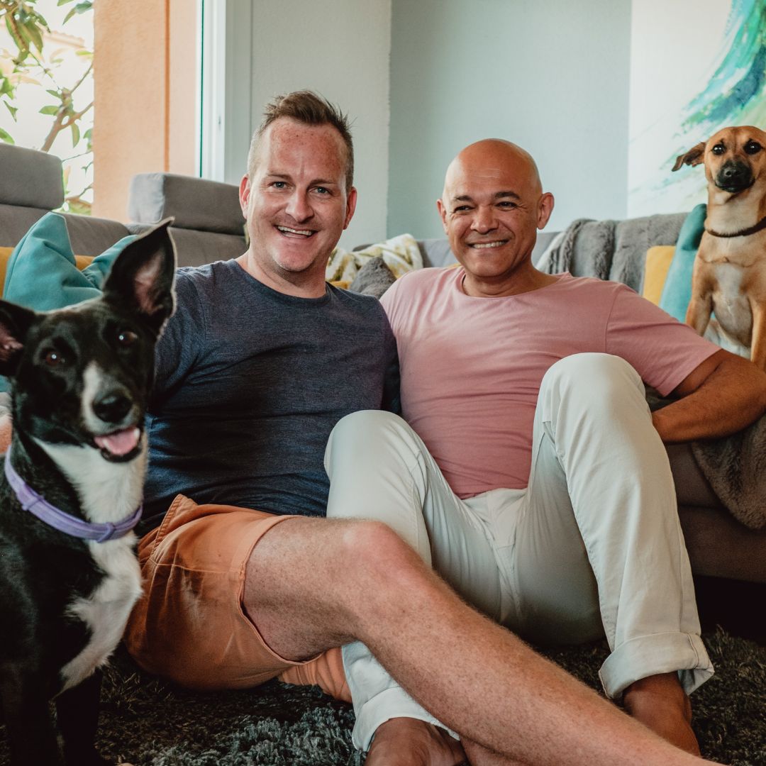 two men sitting on the floor in front of a couch with two dogs
