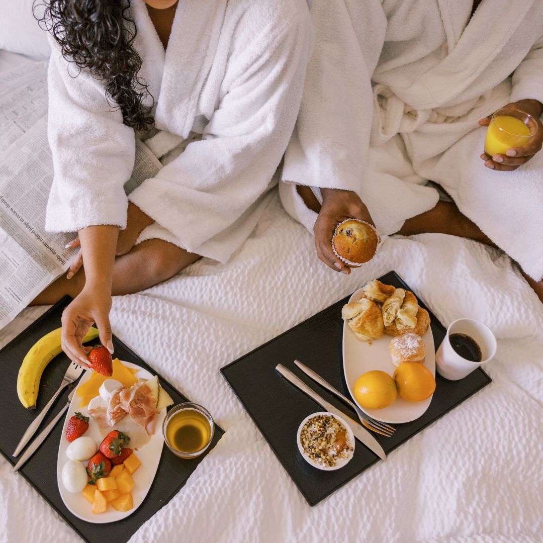 two people in white robes with trays of breakfast in a bed