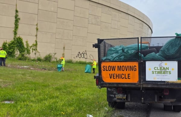 truck filled with garback bags and three people int he distance in brightly colors clothing picking up garbage on the roadside