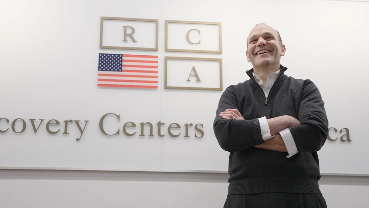 Brett Cohen smiling in front of a wall with Recovery Centers of America logo on it