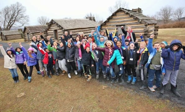 childrean in front of log cabins with their arms in the air
