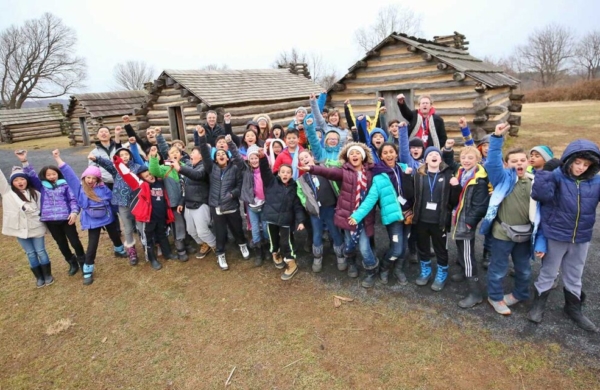 childrean in front of log cabins with their arms in the air