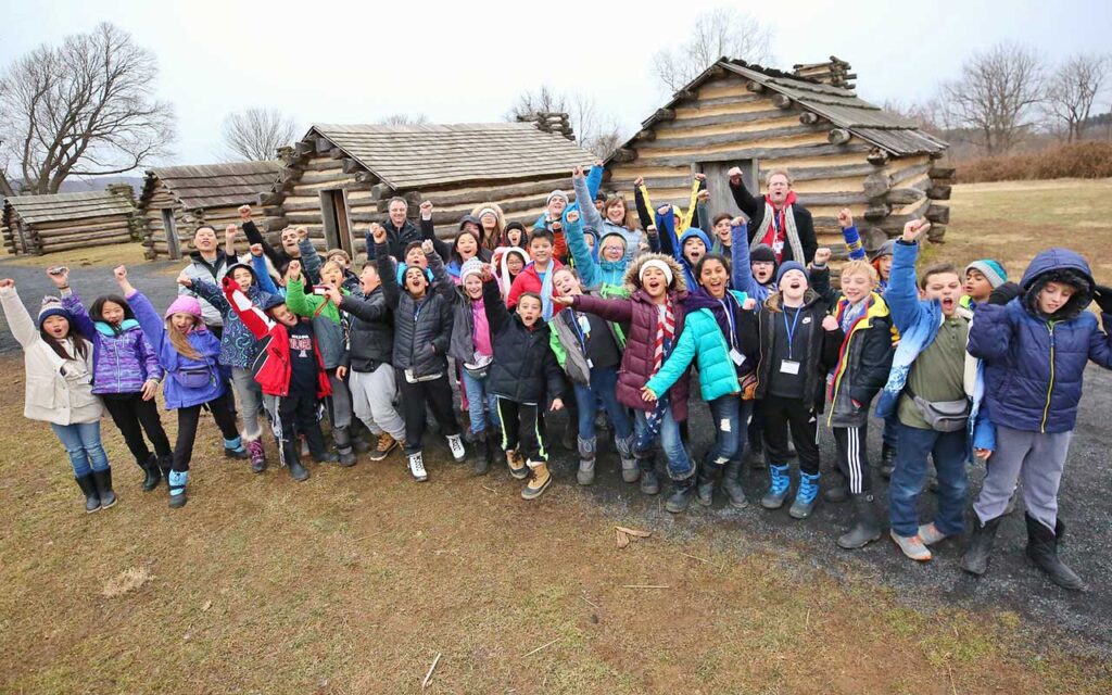 childrean in front of log cabins with their arms in the air