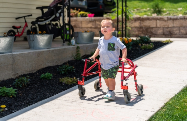 child using a red walking device down an outdoor sidewalk